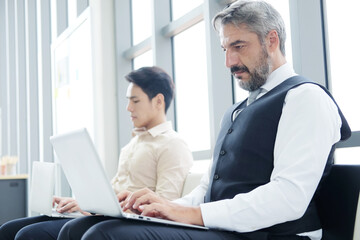 Fototapeta premium Serious Senior Caucasian businessman is working with laptop in workspace at office. He sitting distance from asian staff. Technology for business and New normal lifestyle concept.