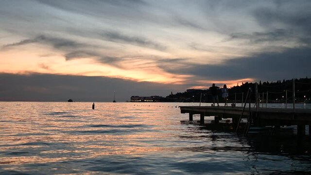 Beautiful sunset golden hour over Portoroz, Slovenia. Beautiful colors reflection on Adriatic Sea water. Summer warm evening. Couple taking photos on concrete pier. Romantic coastal town. Static, wide