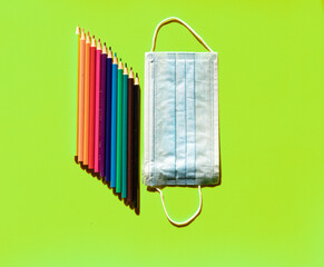 Image of colored pencils, perfectly arranged next to a hygienic mask on a plain green background