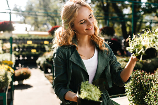 Young Woman In Green Clothes Is Faced With Choosing New Plant For Her Home. Portrait Of Lady With Curls Posing In Botanical Garden