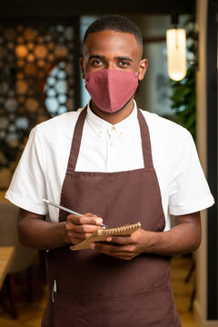 Happy Young African Man In Brown Apron And Protective Mask Taking Orders