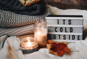 Cozy autumn or winter still life details. Candles and warm sweaters on a wooden background. Light box with phrase It's cold outside. Scandinavian hygge concept.