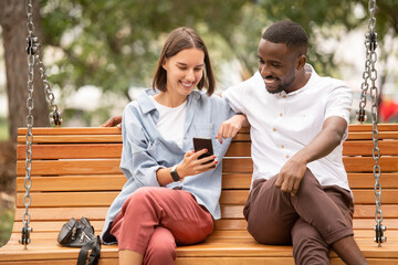 Young smiling woman showing her African boyfriend curious video in smartphone
