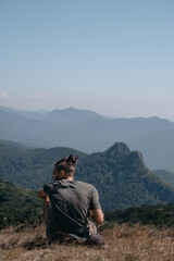 Fototapeta premium Beautiful mountain and hilly landscape in warm, Sunny weather and the young tourist. A male traveler sits on top of a hill and looks at the surrounding mountains.