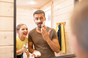 Cute little girl laughing at father applying foam on beard in front of mirror