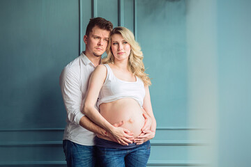 Handsome man and his beautiful pregnant wife are hugging and smiling while standing near the window...