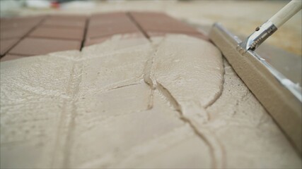 Worker hand with bucket pouring cement for tiles installation making on floor. Road tile with...