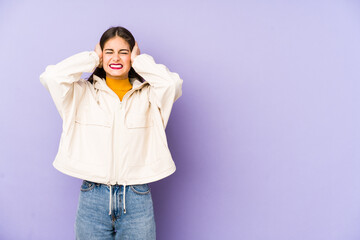 Young caucasian woman isolated on purple background covering ears with hands trying not to hear too loud sound.