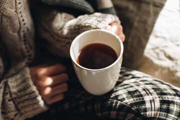 Cozy autumn or winter atmosphere. Young woman with cup of tea and warm sweater is seating at home near the window. Scandinavian hygge concept.