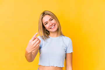 Young blonde caucasian woman isolated on a yellow background pointing with finger at you as if inviting come closer.