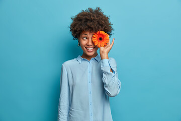 Happy teenage girl has Afro hair and toothy smile covers eye with pretty orange gerbera daisy...