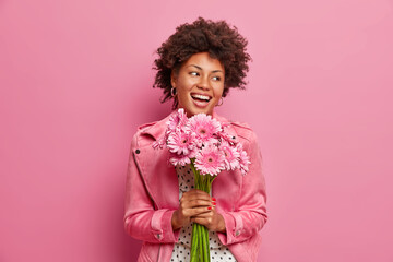 Happy sincere Afro American birthday girl receives flowers from friends, holds bouquet of gerberas,...