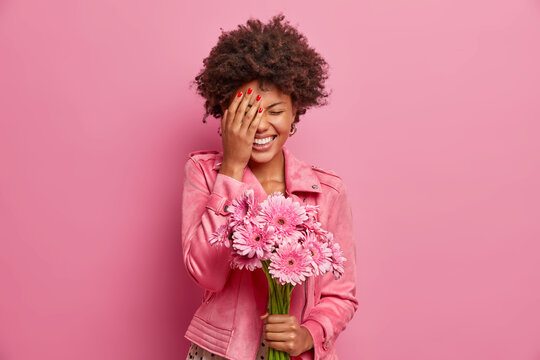 Joyous young African American woman laughs happily, makes face palm, gets nice gift like flowers, holds beautiful gerberas, expresses sincere emotions, isolated on pink background, enjoys smell