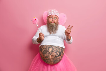 Plump bearded childish male fairy has fun on costume party, dressed in pink pleated skirt, wears wings, has ability to fly, holds magic wand, makes victory gesture, poses against rosy background.