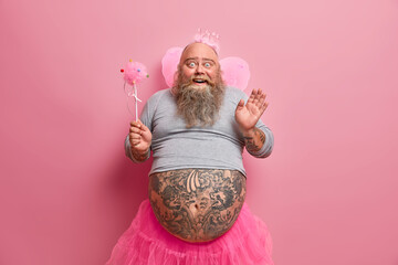 Photo of comic male fairy with thick beard and fat tattooed belly, giggles positively, wears crown...