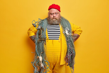 Serious fisherman looks with confident expression, keeps hands on hips, wears striped sailor shirt and yellow overalls, ready to catch fish with net, poses indoor. Fishery and hobby concept.