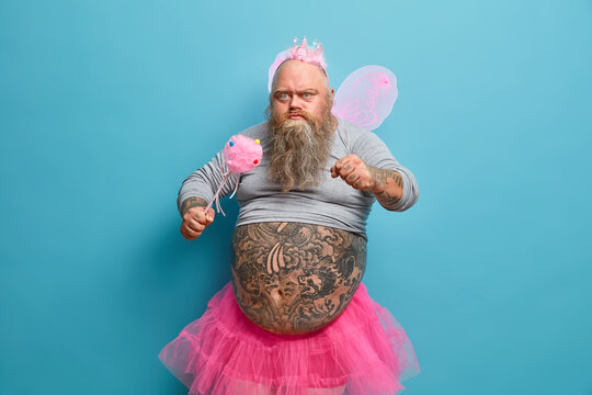 Serious bearded man prepares for fancy birthday party, clenches fist angrily, plays magic fairy, wears butterfly wings, holds magic wand, isolated on blue background. Childrens holiday concept