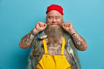 Glad fisherman curls mustache, has thick beard, carries fishing net on shoulders, spends free time for hobby and soul, wears red hat and overalls, has tattoed body, enjoys favourite activity