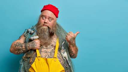 Bearded fisher catches fish with net, enjoys fishing hobby and his occupation, poses with pipe in...