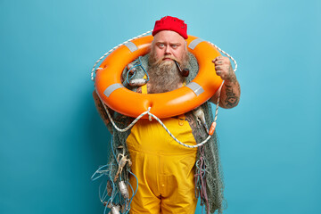 Angry irritaed sailor clenches fist, poses with inflated ring, wears red hat and yellow overalls,...