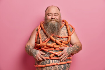 Overeating and excess weight concept. Happy obese man feel satiety after eating sausages, got stout as eats unhealthy junk food, has fat belly, tattooed body, stands with closed eyes, isolated on pink