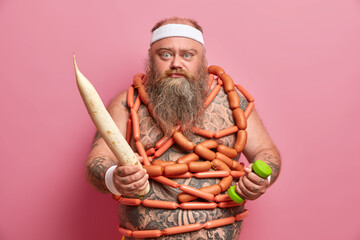 Serious bearded overweight man tries to have healthy nutrition without fast food, wrapped with sausages, holds big fresh white radish, eats low calorie food to lose weight, works with dumbbell