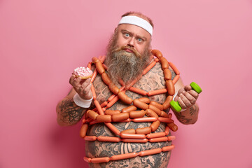 Sad bearded obese man overcomes desire to eat junk food, fights against weight loss, has physical workout, holds dumbbell, wears white headband, has tattooed body, isolated over rosy background