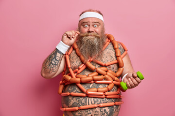Funny surprised fatty guy curls mustache, maintains health, busy doing physical exercises, raises dumbbell, wrapped with sausages, stands indoor. Obesity, workout, dieting, nutrition concept