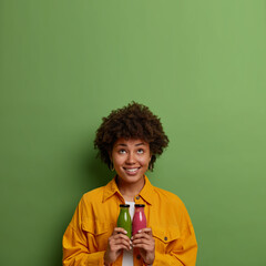 Happy curly haired woman holds glass bottles of freshly made apple and strawberry smoothie, concentrated above with positive expression, has healthy nutrition, copy space on green background.