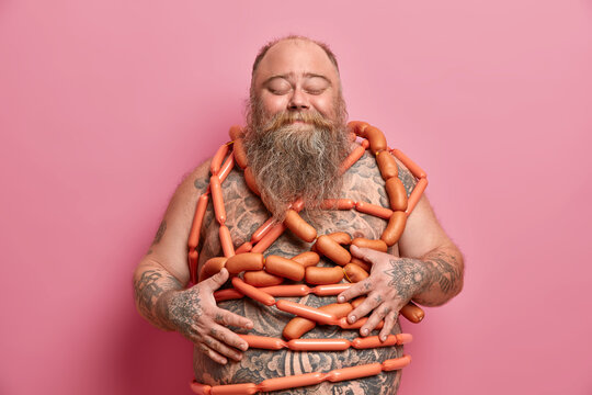 Obese overweight man has unhealthy nutrition, bad habits, wrapped with sausages, has diet failure, feels satiety after eating junk food, regularly overeats, keeps hands on big fat tattooed tummy.