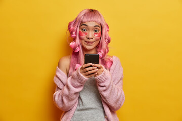 Smiling positive Asian woman holds mobile phone in hands, checks email box, looks gladfully at camera, has pink hair, wears rollers, warm sweater, isolated on yellow background. Technology, beauty