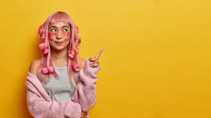 Horizontal shot of pink haired woman points aside on yellow background, advertises product, wears...
