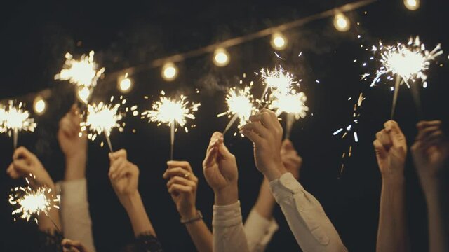 Close-up of hands or palms holding and waving burning sparklers in front of black or dark background. Sparkling lights at birthday party, wedding, New Year, Christmas Eve, Xmas.