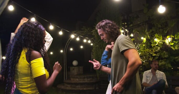 Happy multi-ethnic young people dancing in backyard at party. Portrait of African American girl and Caucasian guy having fun and moving outdoors. Beautiful females and handsome boys dance. Fun concept