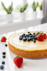 Whole cheesecake with fresh strawberries and blueberries on a white table in white modern kitchen.