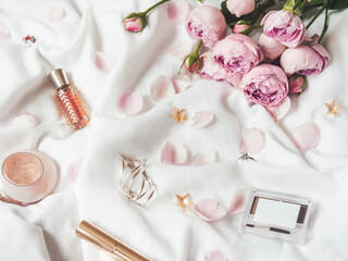 Top view on pink roses and petals on crumpled white fabric background. Decorative cosmetic - perfume, mascara, eye and brow shadows. Cute and fashionable jewelry accessories.