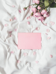 Roses and petals on crumpled white fabric. Natural elegant decoration. Romantic background with copy space on pink envelope. Top view, flat lay.