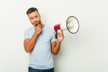 Young mixed race asian man holding a megaphone looking sideways with doubtful and skeptical expression.