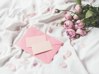 Roses and petals on crumpled white fabric. Natural elegant decoration. Romantic background with copy space on pink envelope and visit cards. Flat lay.
