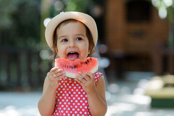 Little 2 year old girl kid in red dress with white polka dots and hat is eating a very funny slice of a watermelon.