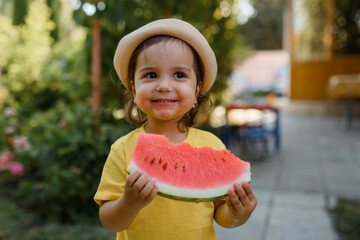 Little 2 year old girl kid in  yellow t-shirt and hat is eating a very funny slice of a watermelon.