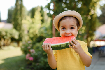 Little 2 year old girl kid in  yellow t-shirt and hat is eating a very funny slice of a watermelon.