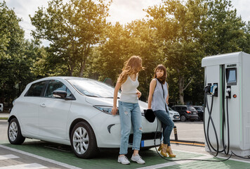 Fototapeta Two happy young beautiful women are talking to each other until their electric car is charging at the charging station situated in the car park. obraz