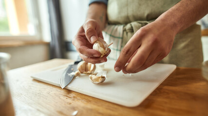 Close up of hands of man in apron peeling garlic while preparing a meal in the kitchen. Cooking at...
