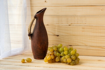 Still life with green grapes and an earthenware jug. Grapes and a jug on a light natural wooden background. Rustic style. Warm vintage flavor. Autumn mood. The concept of winemaking.