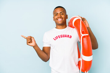 Young latin life guard isolated on blue background smiling and pointing aside, showing something at...