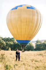 Obraz premium Full length portrait of lovely young couple in black, hugging each other and enjoying summer walk in the field, waiting for their air balloon tour. Yellow air balloon on the background