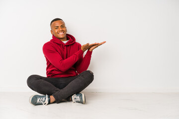 Young latin man sitting on the floor isolated holding a copy space on a palm.