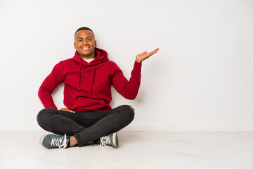Young latin man sitting on the floor isolated showing a copy space on a palm and holding another hand on waist.