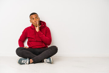 Young latin man sitting on the floor isolated is saying a secret hot braking news and looking aside